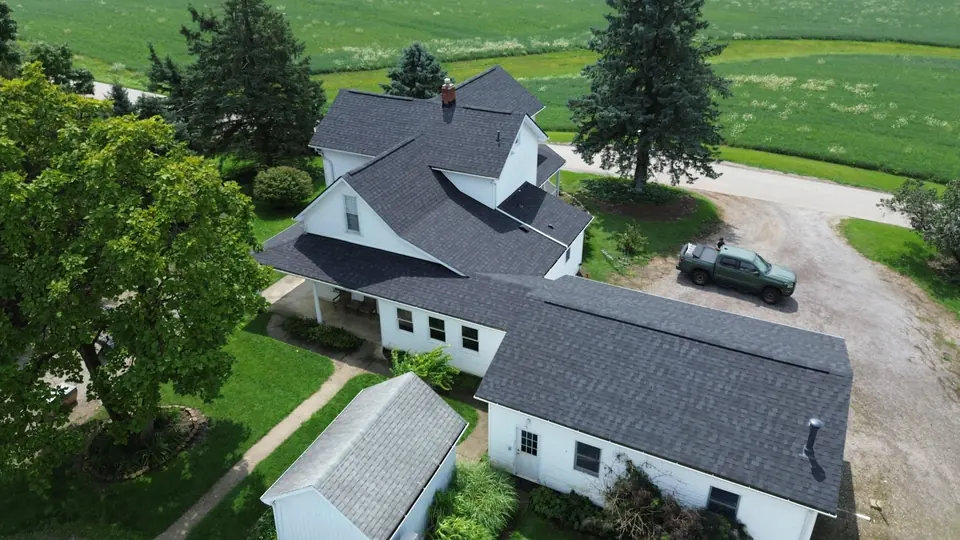 Aerial view of a farmhouse with a new dark shingle roof, alongside a garage and outbuilding.