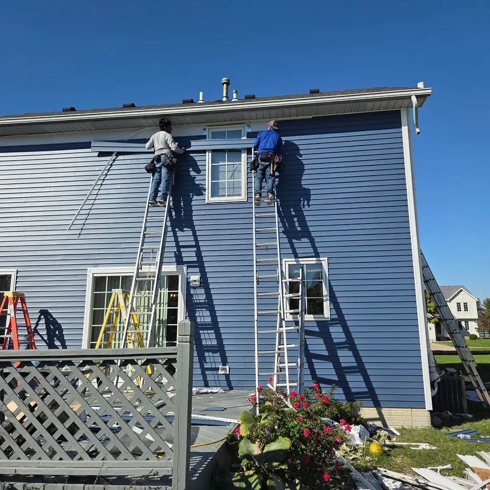 Two installers fitting blue-gray siding and trim from ladders on the side of a two-story home.
